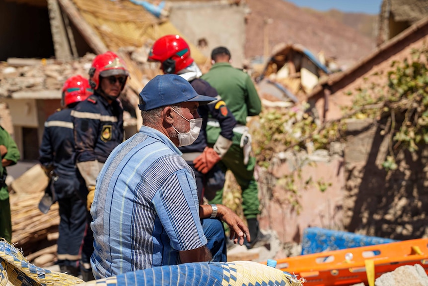 A man wearing a blue cap and striped shirt sits down and watches rescue workers clear rubble.