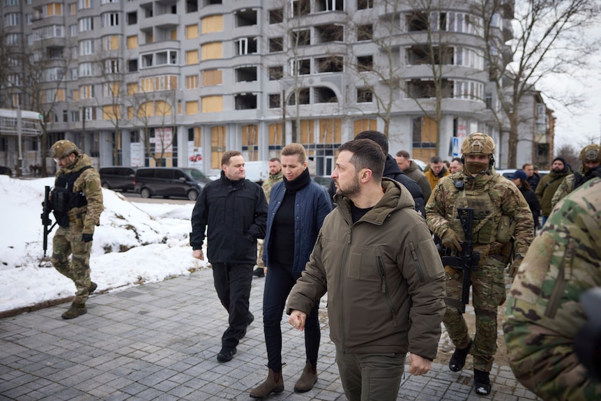 Politicians are escorted by armed soldiers as they walk among buildings past snowy ground.