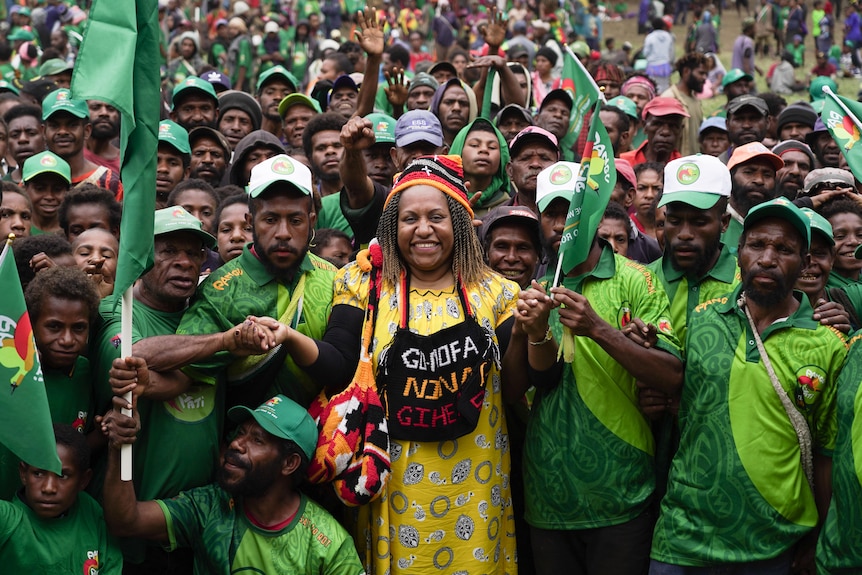 A woman in a yellow dress smiles at the centre of a large crowd dressed in green and waving flags