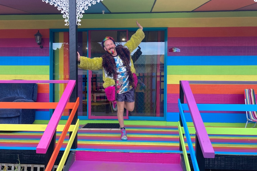 A man with rainbow-coloured hair and dressed in bright clothes stands outside a rainbow-coloured house.