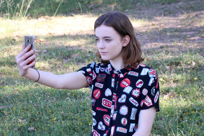 Charlotte sits on the grass, holding her phone up in front of her to take a selfie.