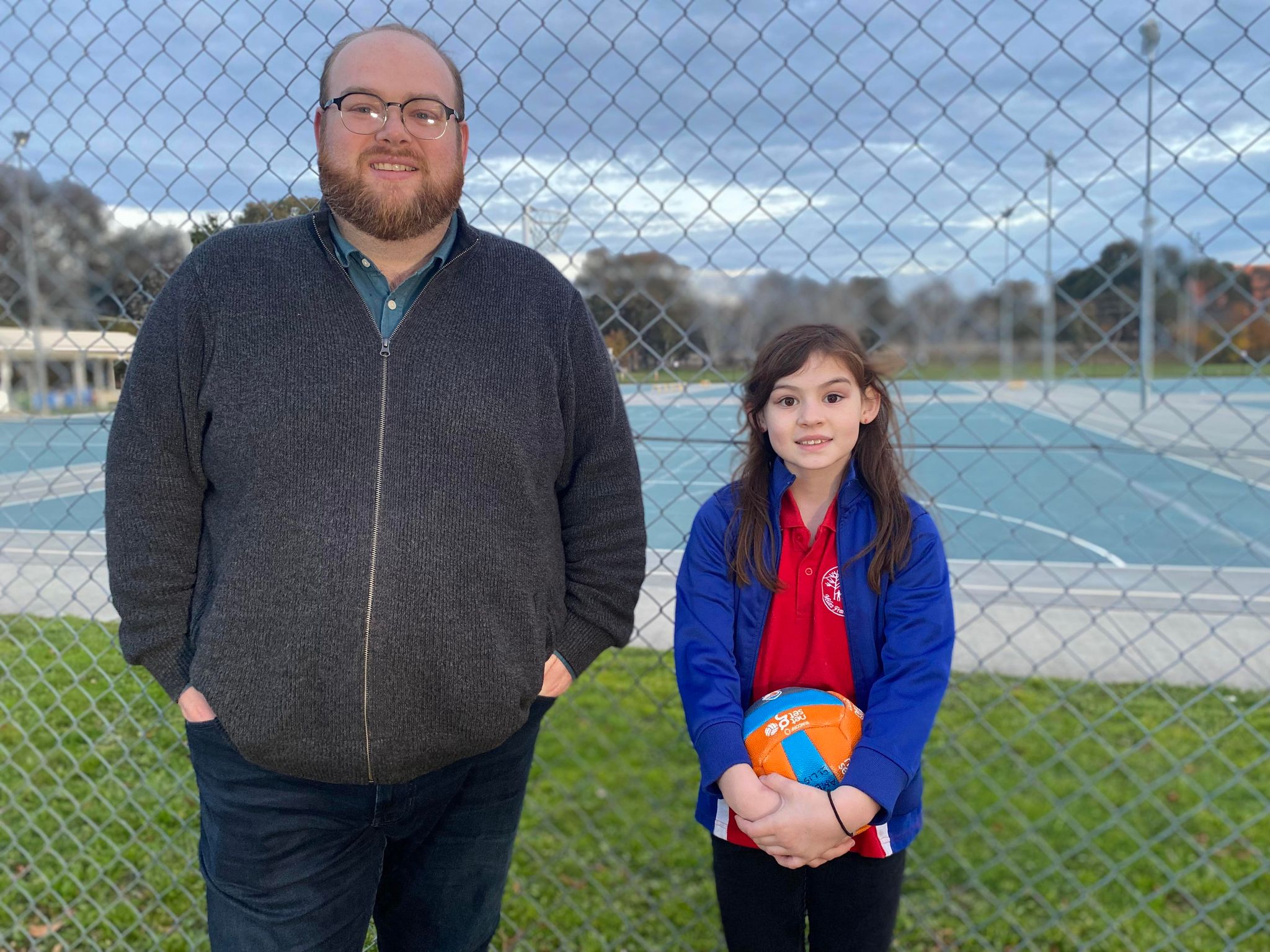 A father and daughter stand out the front of netball courts, the daughter is holding a netball. 