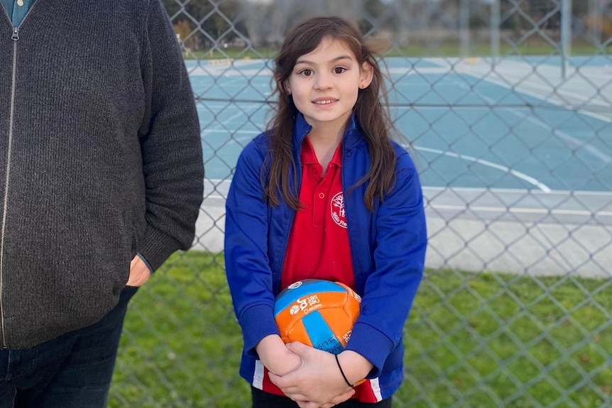 A father and daughter stand out the front of netball courts, the daughter is holding a netball. 