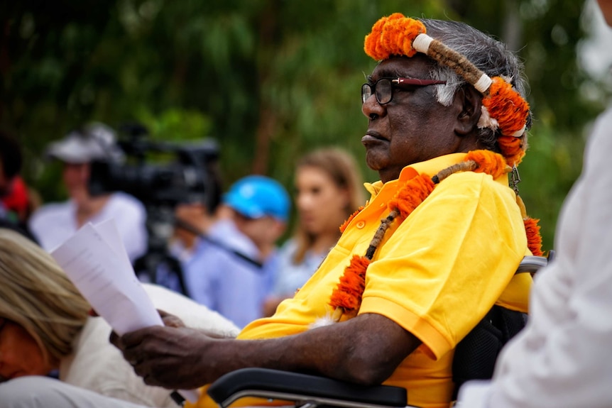 Galarrwuy Yunupingu sits in a wheelchair wearing a yellow shirt and traditional headdress.