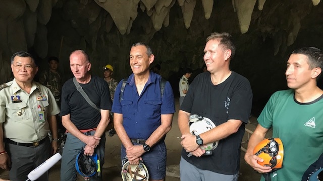 A group of five men stand in a cave with stalactites above their heads seemingly answering journalists' questions