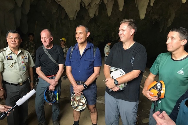 A group of five men stand in a cave with stalactites above their heads seemingly answering journalists' questions