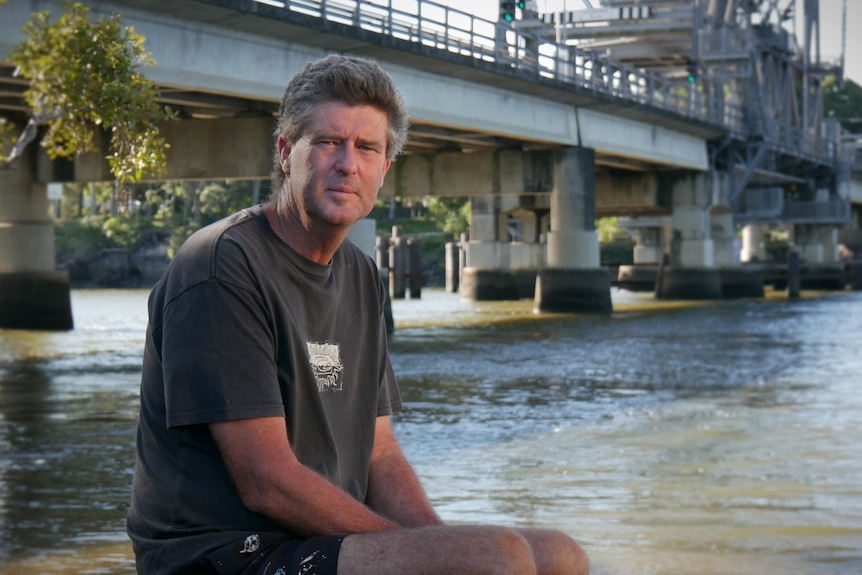 A man in a grey shirt sitting in front of a river with a big concrete bridge behind.