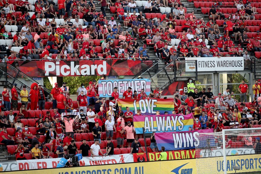 A crowd wearing red holds up rainbow pride banners in the stands during a game