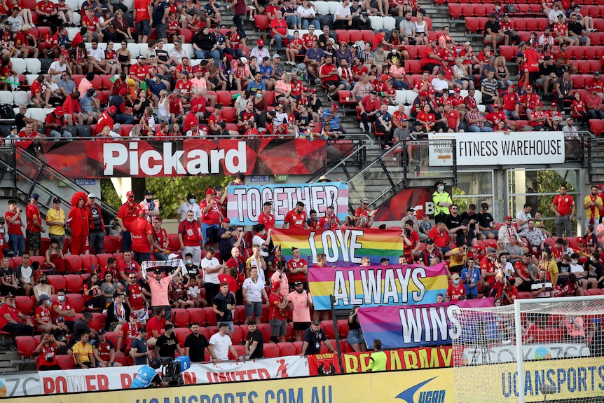 A crowd wearing red holds up rainbow pride banners in the stands during a game