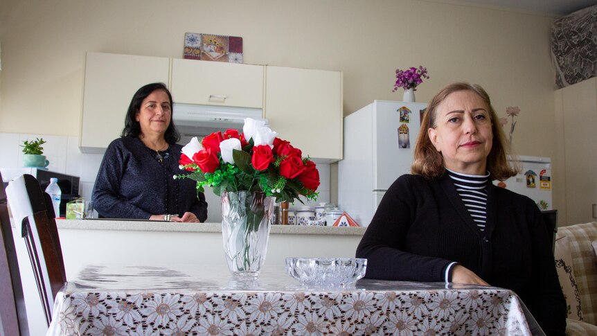 One woman stands at the kitchen bench, the other sits at the table looking at camera