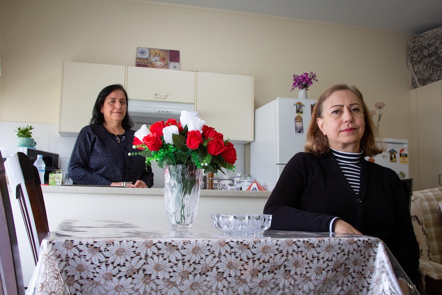 One woman stands at the kitchen bench, the other sits at the table looking at camera