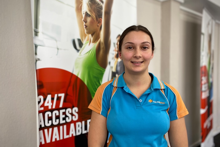 A young woman with brown hair wearing a blue/orange shirt stands next in front of a fitness poster with a woman exercising
