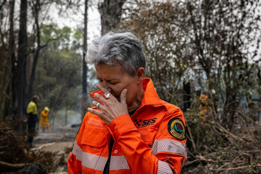 An SES volunteer stands in front of the remains of a bushfire, her hand to her face in sorrow.