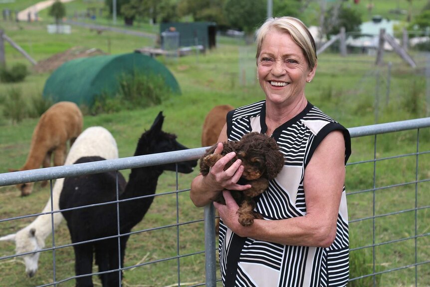 Sue Barker standing on a property holding a puppy and smiling. There are alpacas in the paddock behind her.