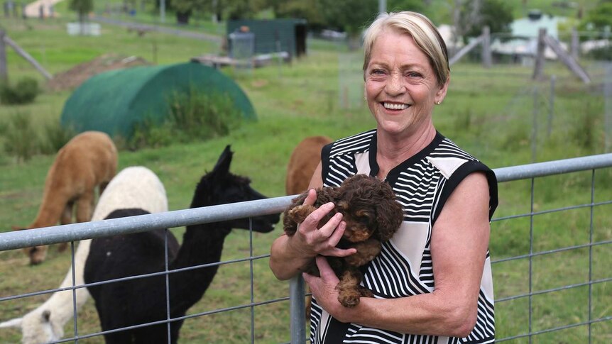 Sue Barker standing on a property holding a puppy and smiling. There are alpacas in the paddock behind her.