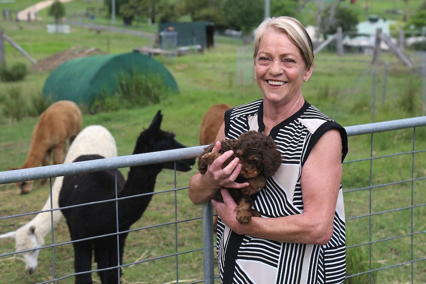Sue Barker standing on a property holding a puppy and smiling. There are alpacas in the paddock behind her.
