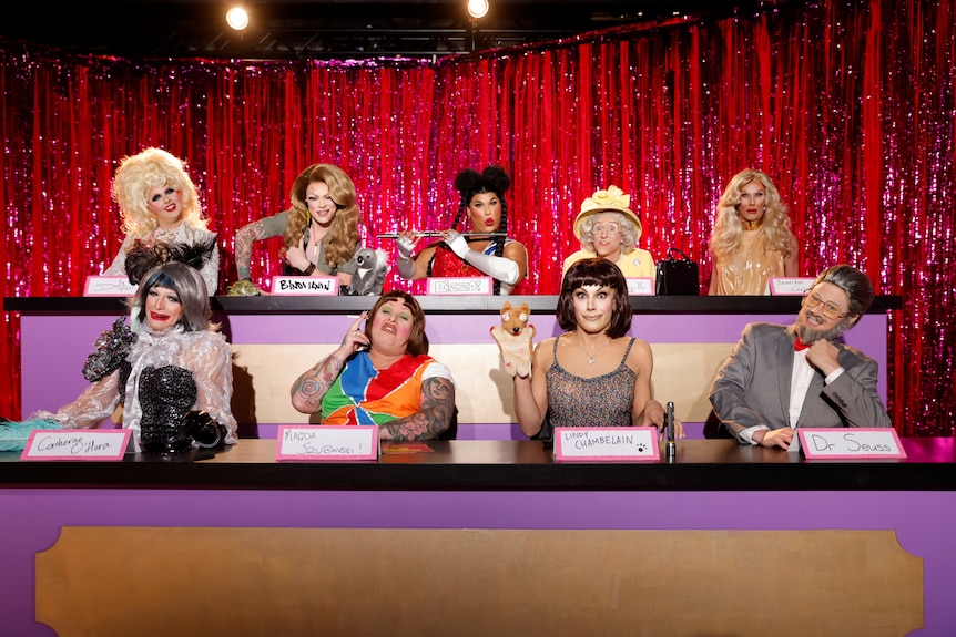 A group of drag queens dressed as different characters sitting in two rows in Snatch Game in RuPaul's Drag Race Down Under