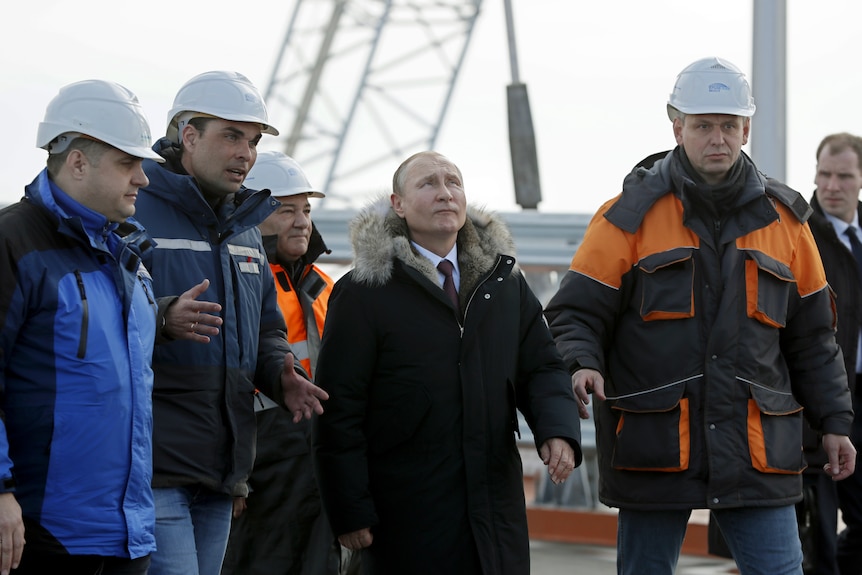 Vladimir Putin, centre, inspects the road section of a bridge. He looks up. Four men wearing hard hats surround him. 