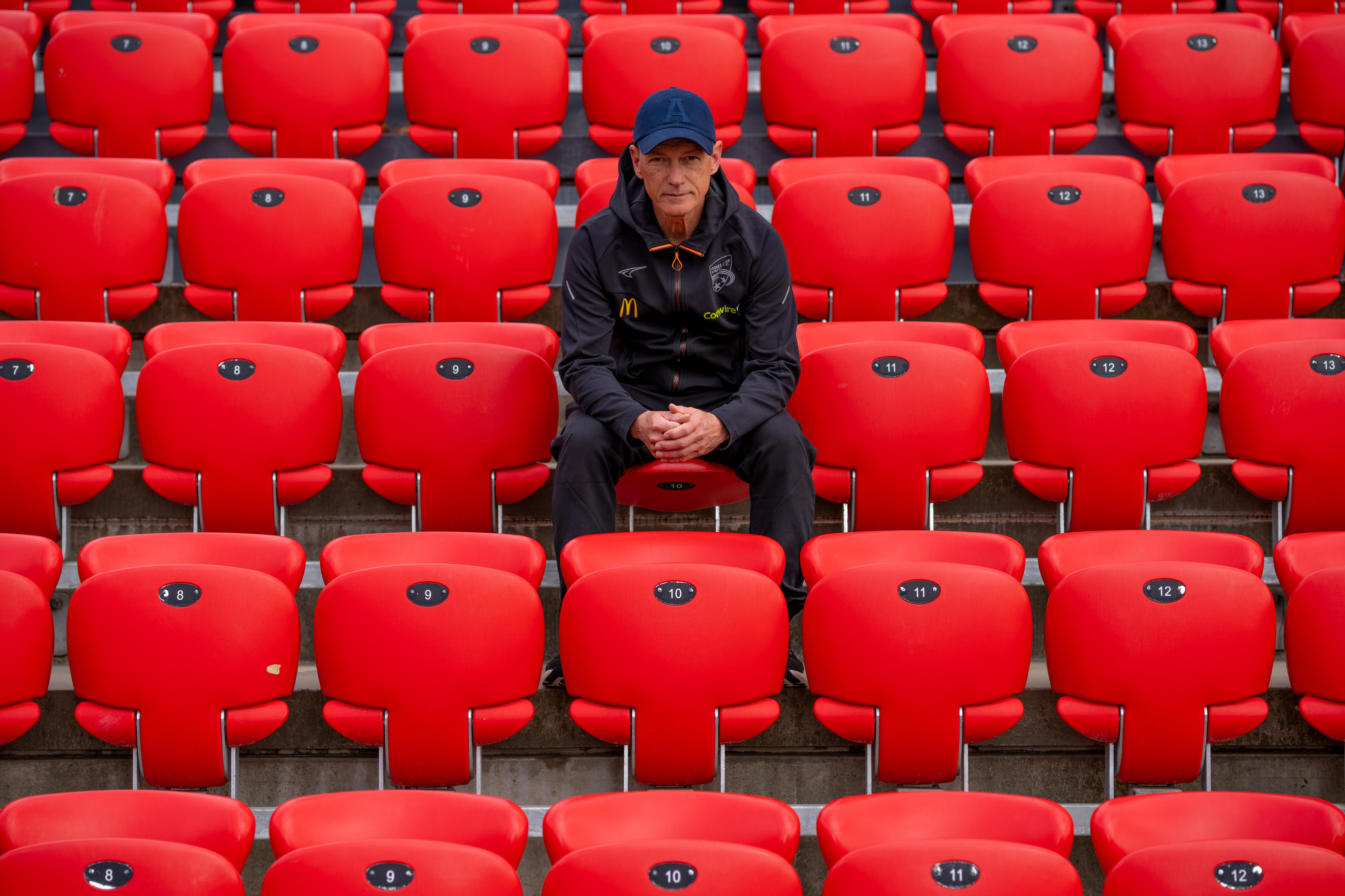 A man in a cap sits in the audience seats overlooking a soccer field with AUFC emblazoned in the audience stand