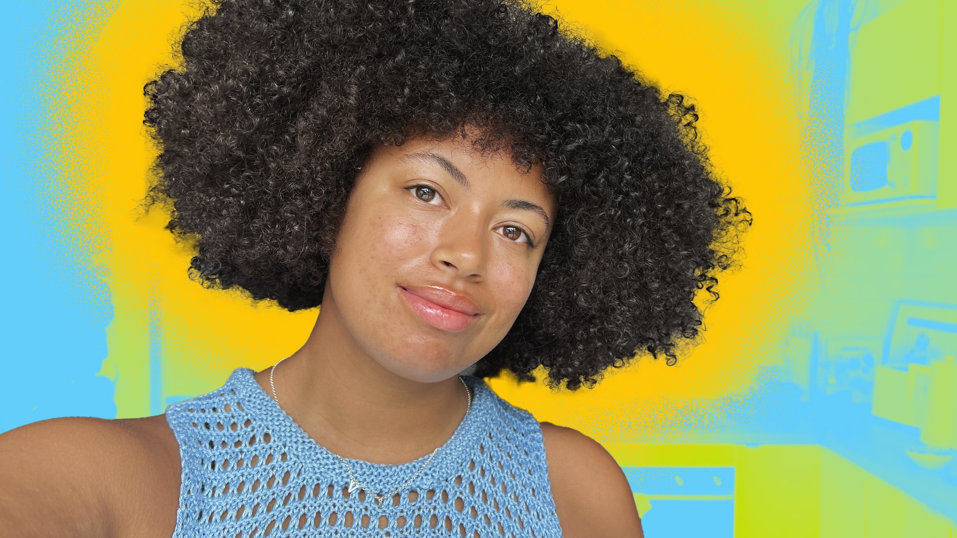 Yasmin smiles as she looks to the camera in a selfie, with her short afro out, standing in a kitchen coloured blue and yellow.