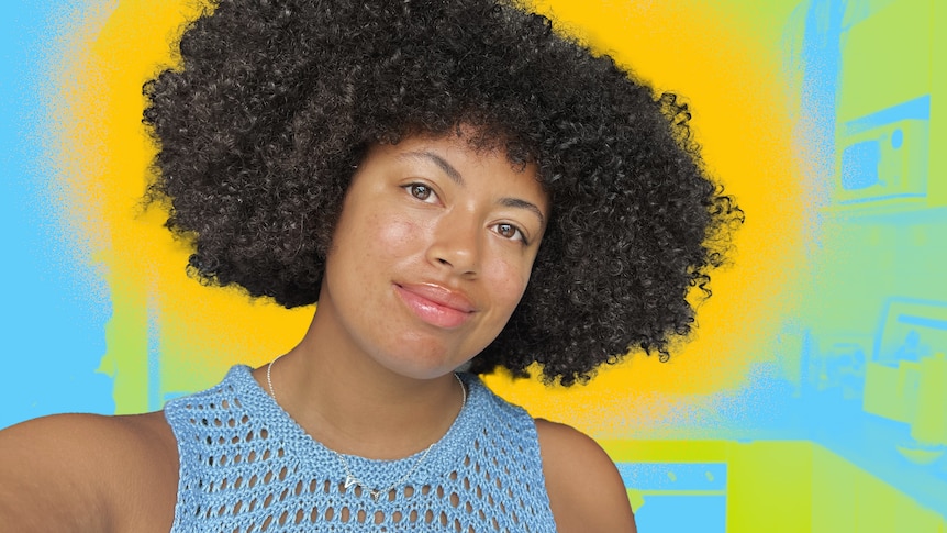 Yasmin smiles as she looks to the camera in a selfie, with her short afro out, standing in a kitchen coloured blue and yellow.