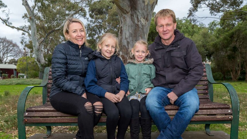 The Squires family, Danielle, Olivia, Zoe and Daryl sit on a park bench and smile at the camera.