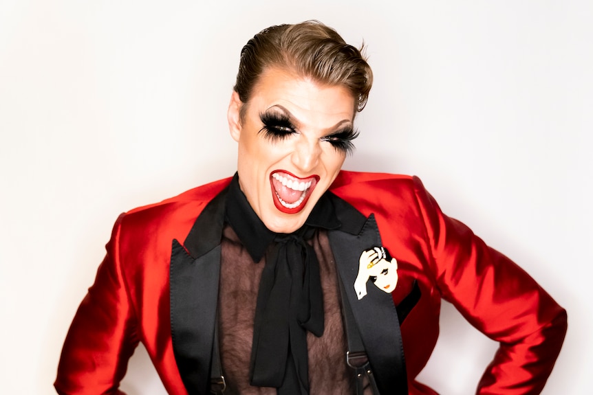 A man in his 30s in drag makeup, in a black sheer shirt and shiny red jacket, open wide smiling mouth