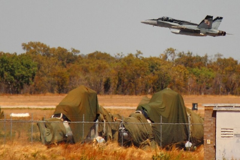 Two choppers are behind a fence and covered with a sheet. A RAAF plane flies overhead.