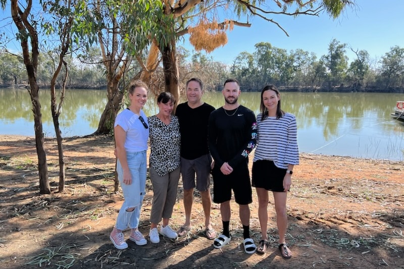 A family of five people standing on the banks of the River Murray. The river reflects the blue sky and trees.