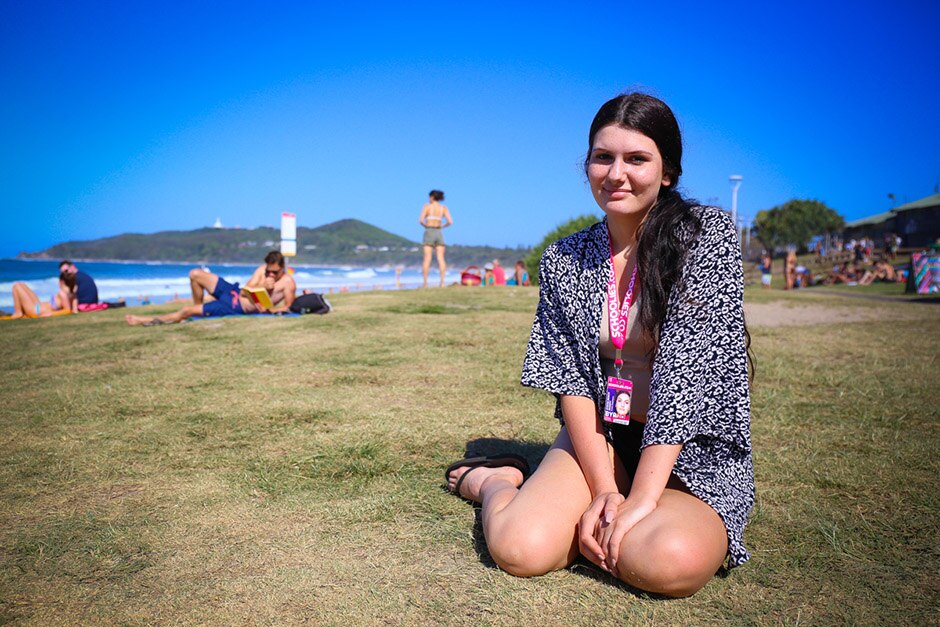 A young woman sitting cross-legged on grass, with a beach and other beach-goers in the background.