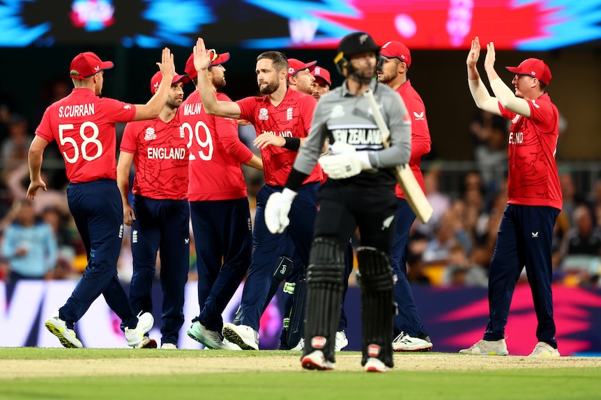 A group of England cricketers celebrate in the background as a New Zealand batsman walks off toward the camera.