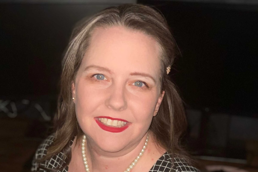 Sarah Taylor, wearing a white necklace, sits on a chair and smiles for the camera.