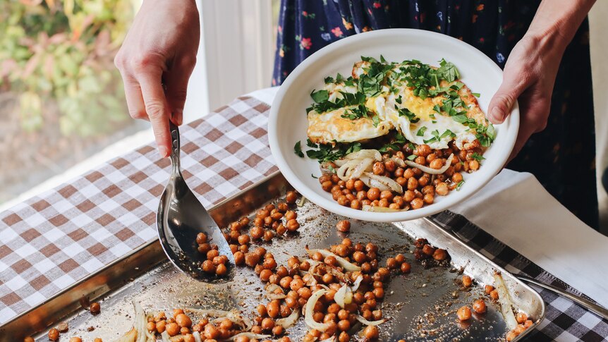 A pair of hands spoons roasted chickpeas from a tray into a dish with eggs and herbs.