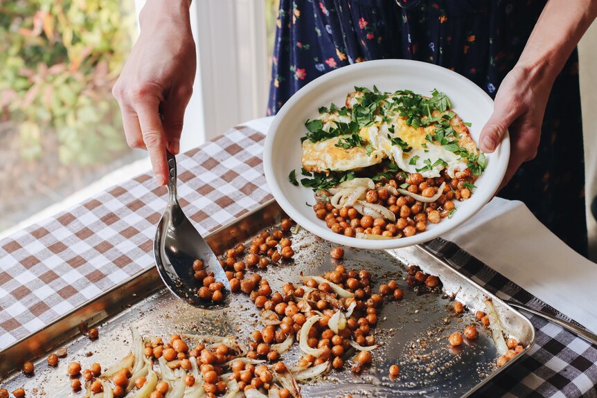 A pair of hands spoons roasted chickpeas from a tray into a dish with eggs and herbs.