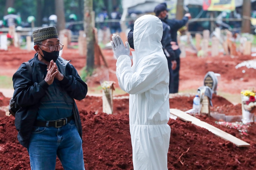 A man in full PPE clasps his hands in prayer to an Indonesian man in a face mask while they stand in a graveyard