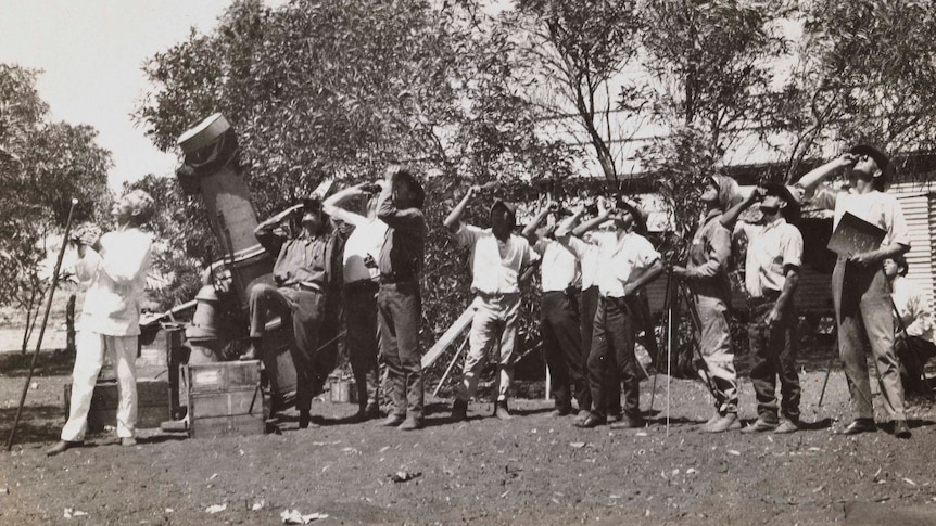 A sepia photo of men looking up, holding shade over their eyes, and a telescope