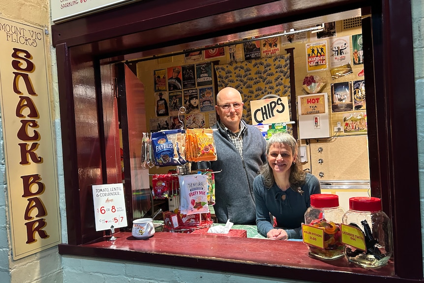 A man and a woman at a counter with lolly packets on a stand. 
