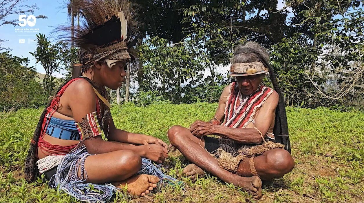 A young girl sits in green outdoors with her grandmother, she teaches her how to weave. 