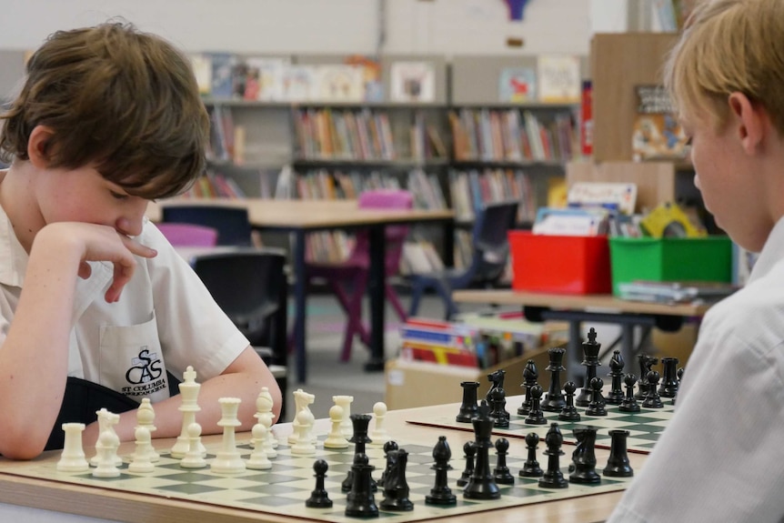 Two primary school students sit at a chess board, looking at the pieces.