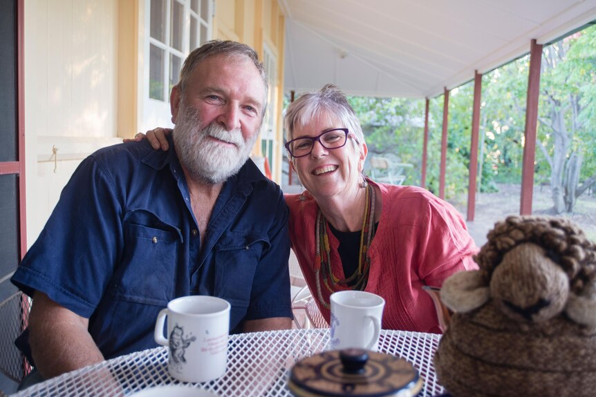 An older bearded man and his smiling wife sit at a table with coffee cups in front of them.