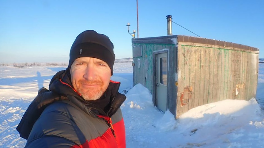 A man in a coat and beanie standing in the snow in front of a small shack.