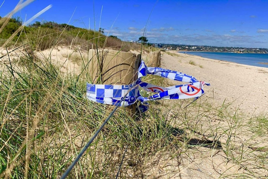 Blue and white police tape is wrapped around a timber post on the edge of the sand dunes on Seaford beach.