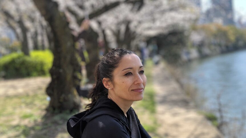 A woman looks out to a river as she sits in front of a row of cherry blossoms.