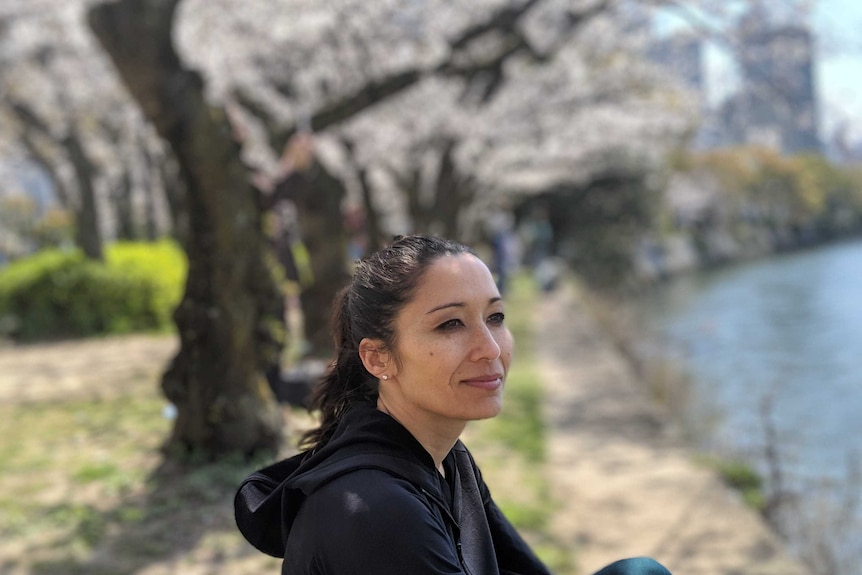 Kumi Taguchi looks out to a river as she sits in front of a row of cherry blossoms.