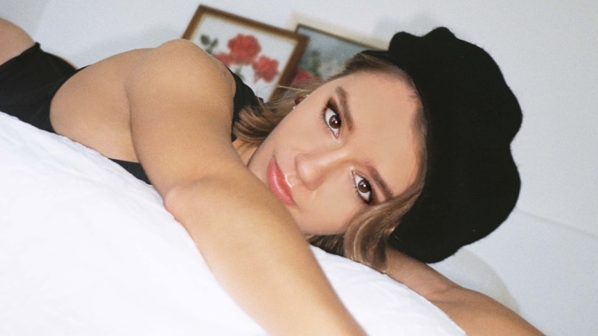 Image of Eves Karydas, wearing black hat and outfit, laying on a bed