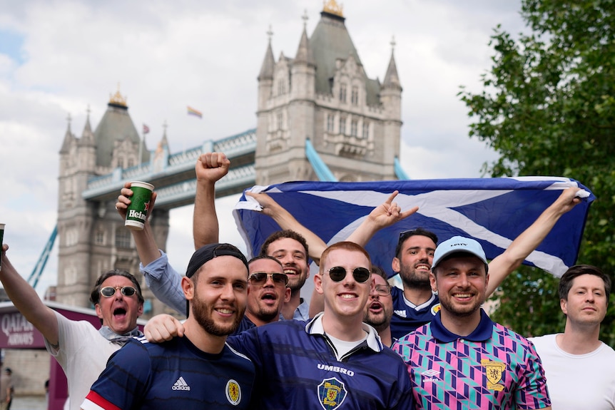 A group of men wearing blue and holding Scottish flags stand and smile, with Tower Bridge in the background