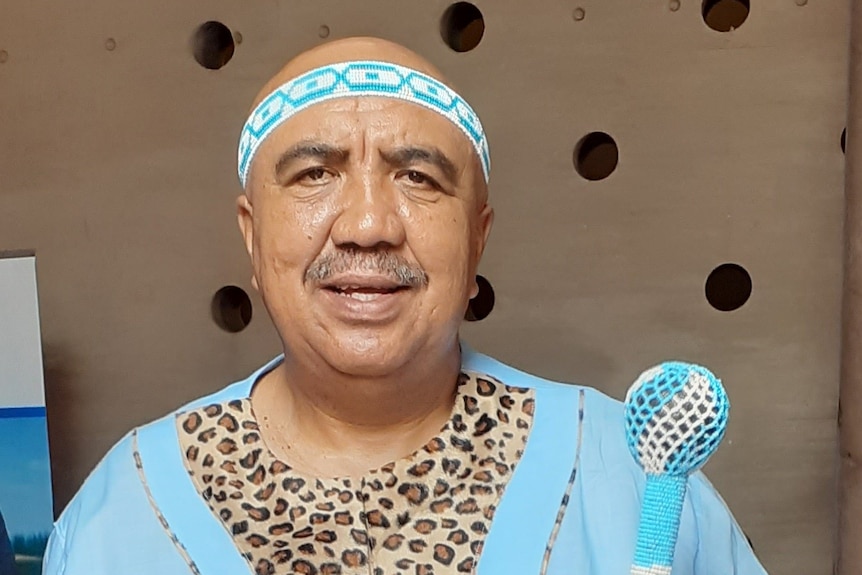 Glen Taaibosch, in long blue shirt with blue headband, holding a rod or stick, smiling.