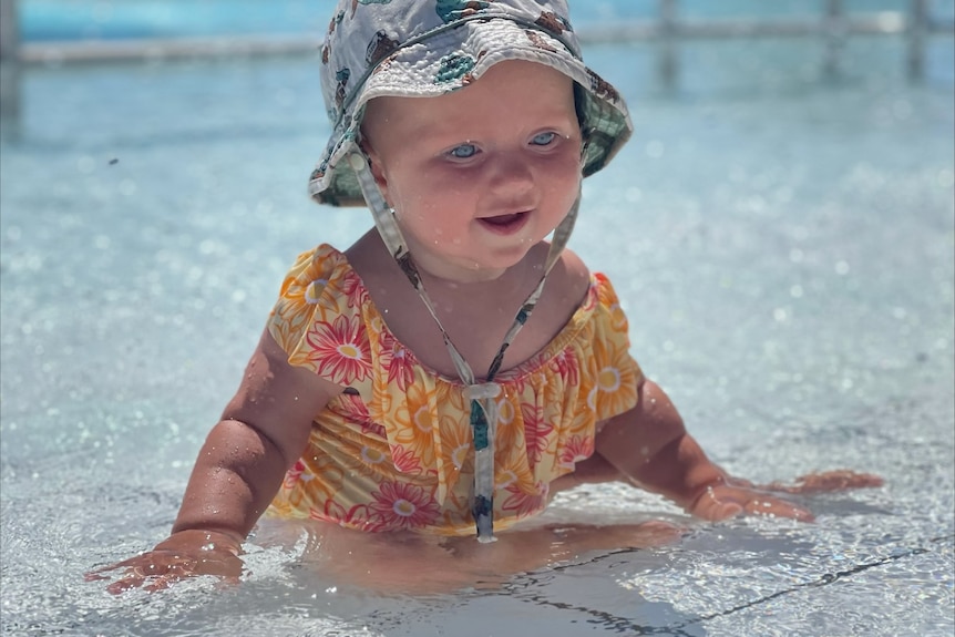 baby with hat on and swimming suit, sitting in shallow water 