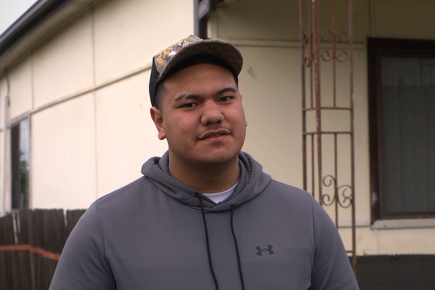 A man wearing a hoodie and cap looks at the camera standing in front of a fibro low-set house.