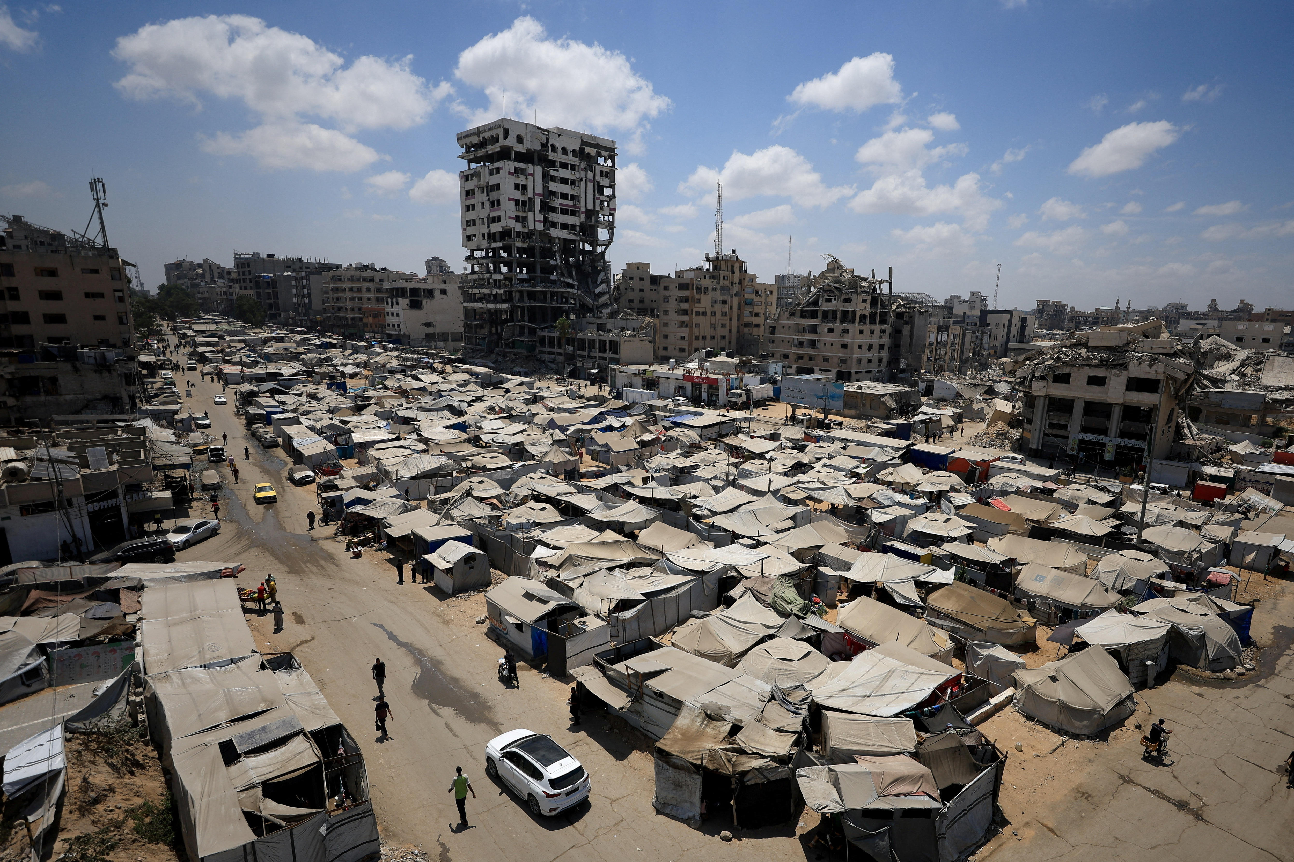 dozens of make shift tents on dirt land with destroyed buildings behind them.